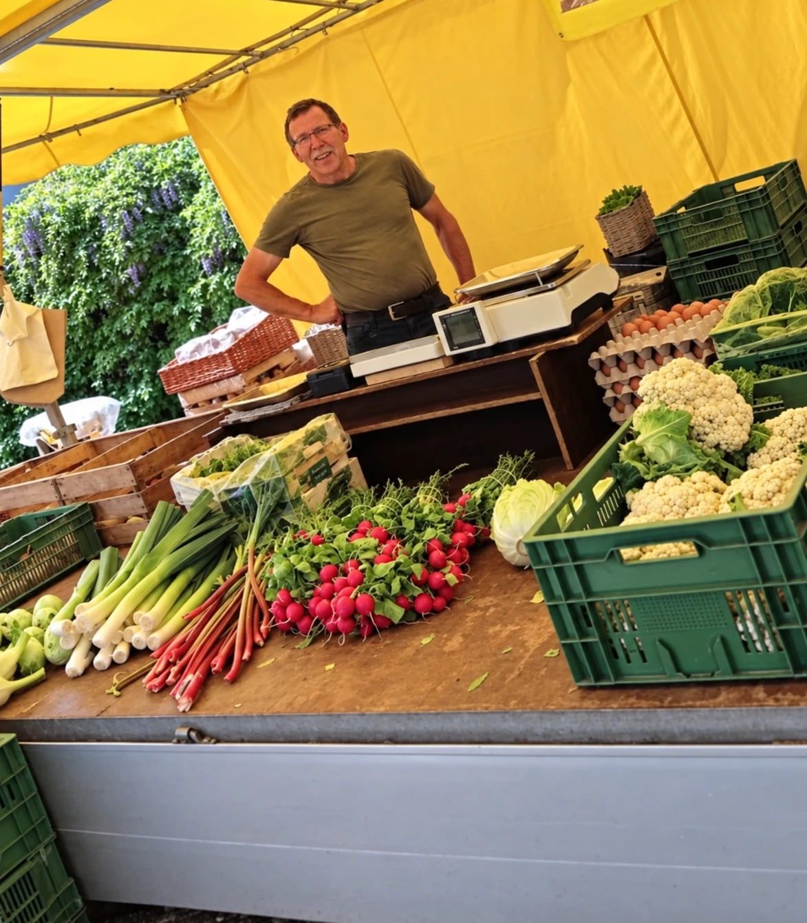 Nobbi hinter seinem Marktstand unter gelbem Dach, davor Lauch, Radieschen, Rhabarber, Blumenkohl und Eier.