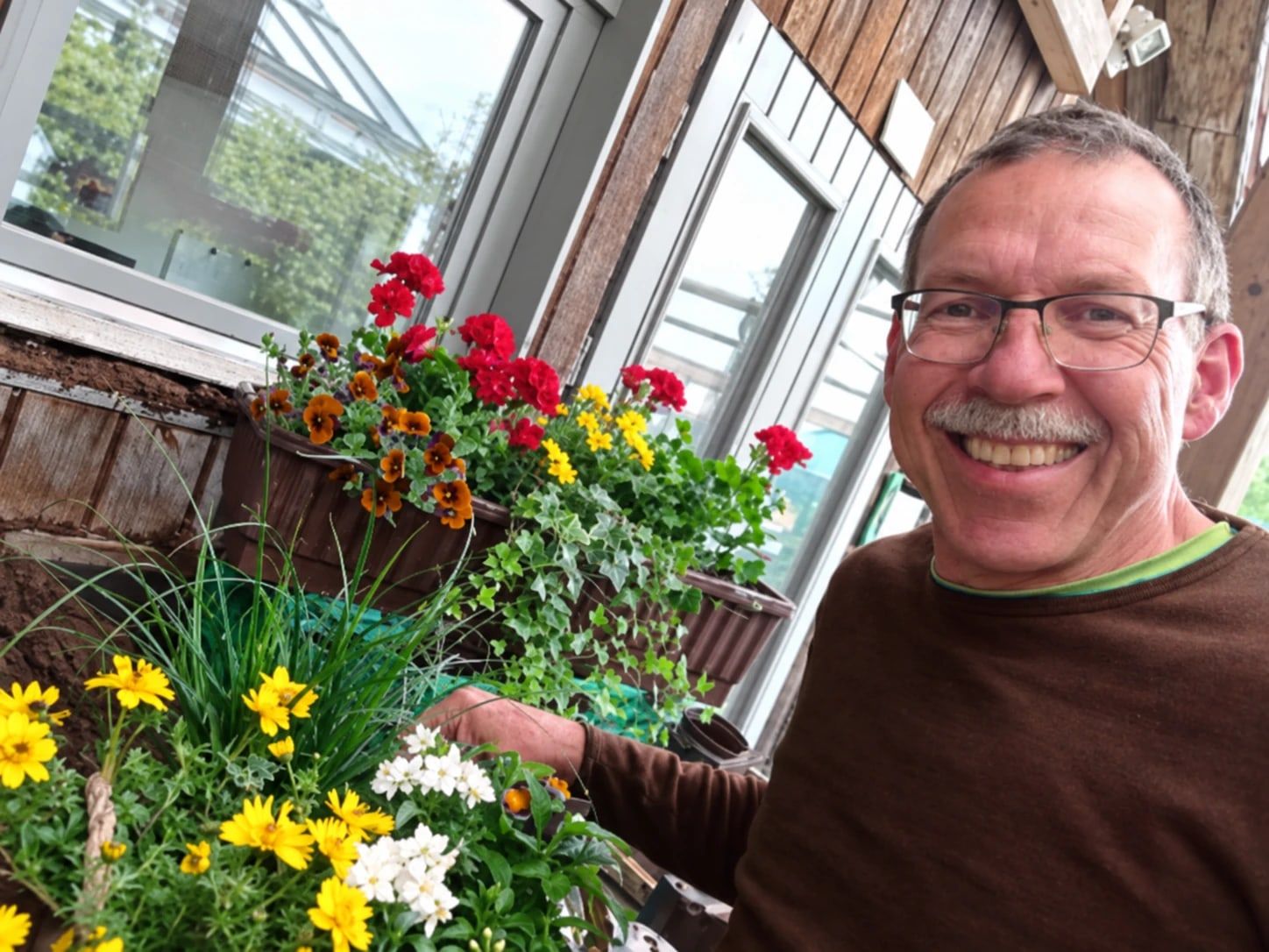 Nobbi lächelt zwischen Blumenkästen mit Geranien, Hornveilchen und Margeriten vor dem Gewächshaus.